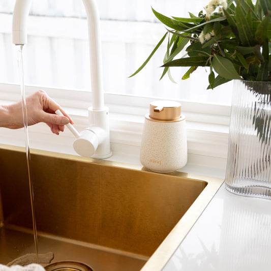 Ceramic soap dispenser bottle on a gold kitchen sink with a hand on a white tap